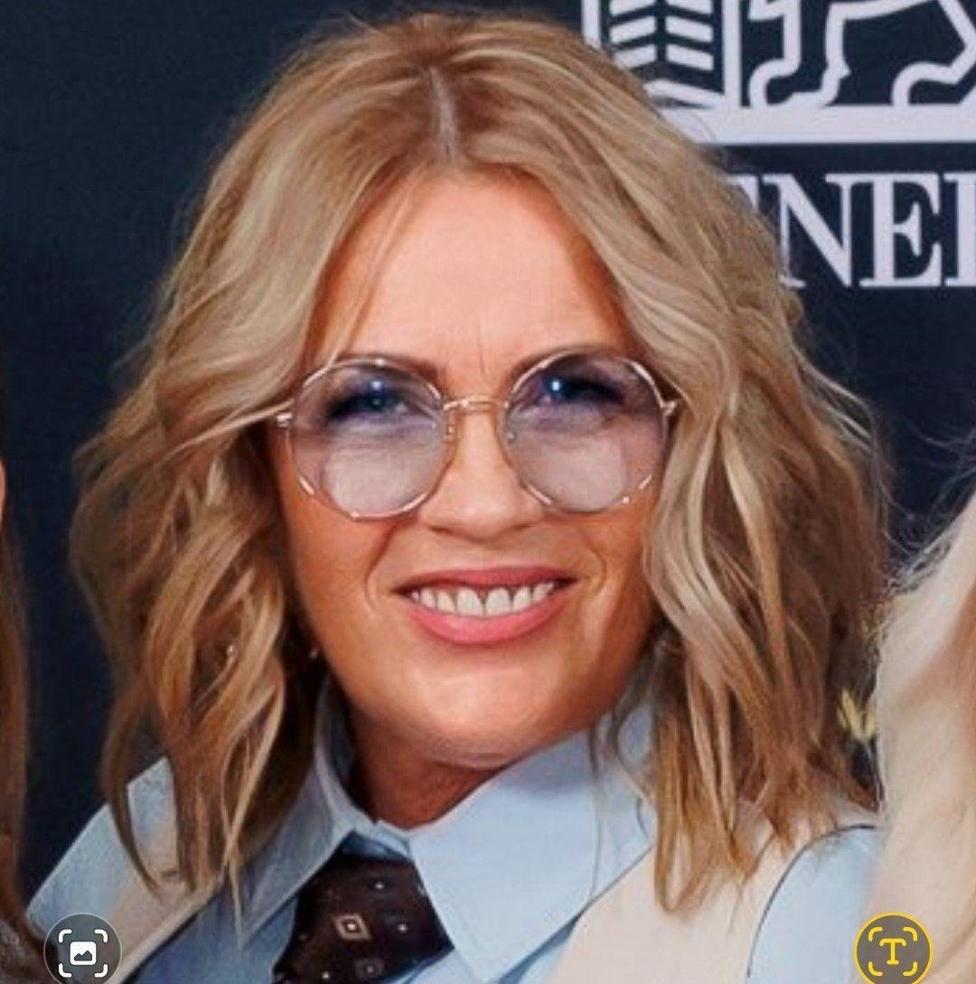 Portret uśmiechniętej kobiety w okularach, blond włosach i koszuli z krawatem, na tle ciemnego tła z logotypem. Kadr od ramion w górę, z widocznym fragmentem drugiej osoby.