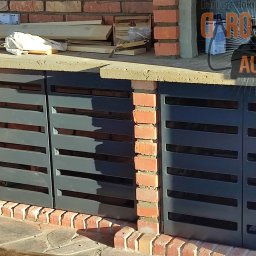 DARO-SPAW AUG DARIUSZ HOŁUBOWICZ - Zabudowa grilla z metalowymi, poziomymi lamelami w kolorze grafitowym, osadzona w ceglanej konstrukcji, widoczne narzędzia i materiały budowlane na blacie.