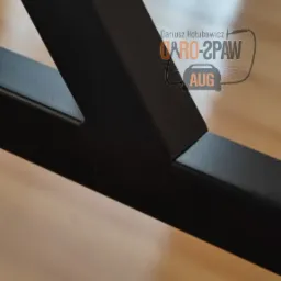 Szczegółowe ujęcie spawu na czarnej, metalowej konstrukcji, widoczny fragment logo firmy w tle, na drewnianej podłodze.