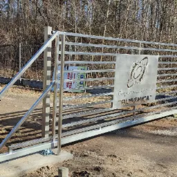 Przesuwna brama wjazdowa z metalowymi poprzeczkami i ozdobnym panelem z wyciętym logo Gospodarstwa Rybackiego 'Bliznica', widoczny mechanizm jezdny i tło z drzewami.