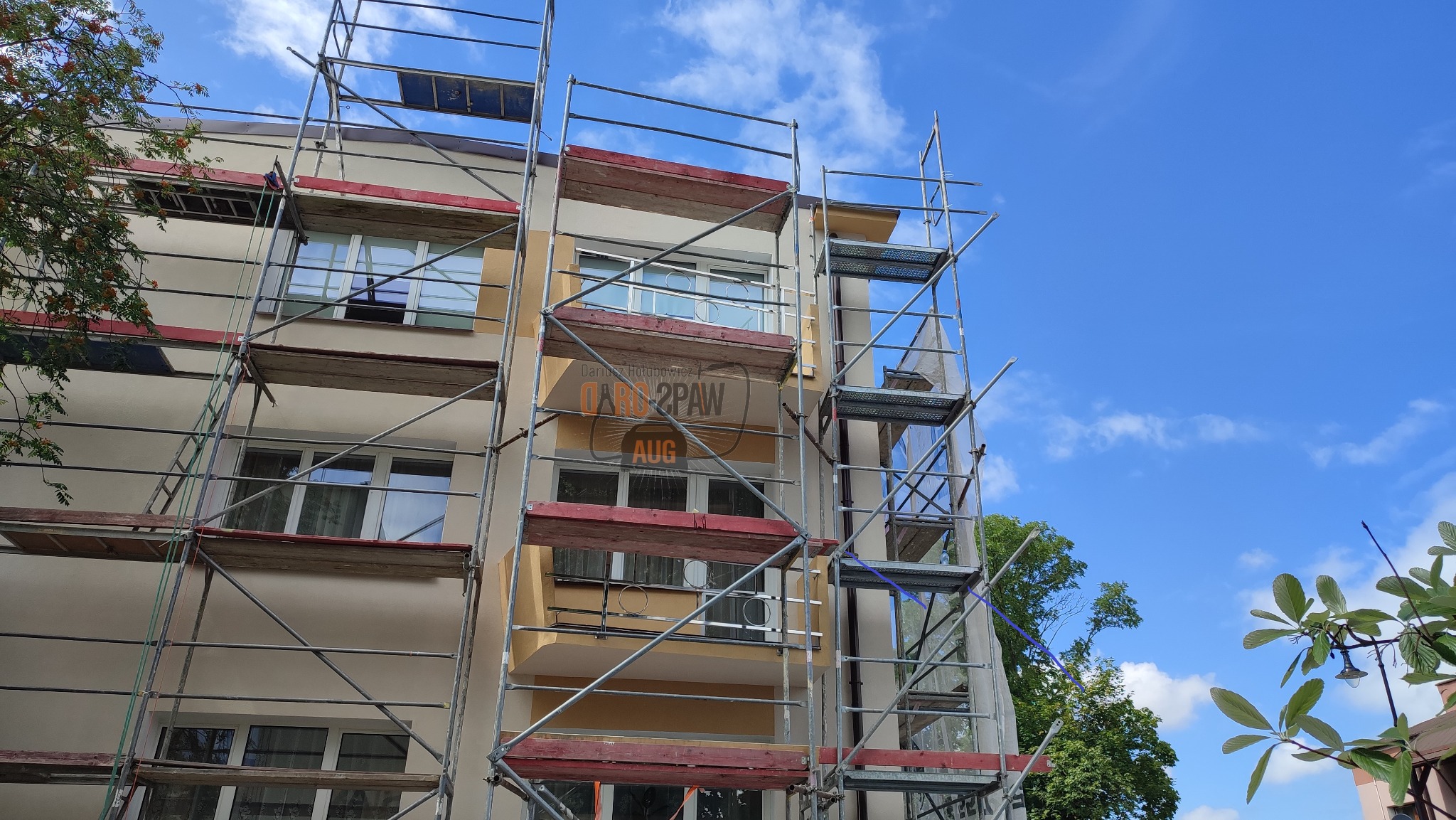 Budynek wielorodzinny w trakcie remontu elewacji, otoczony metalowymi rusztowaniami, widok na błękitne niebo z chmurami.
