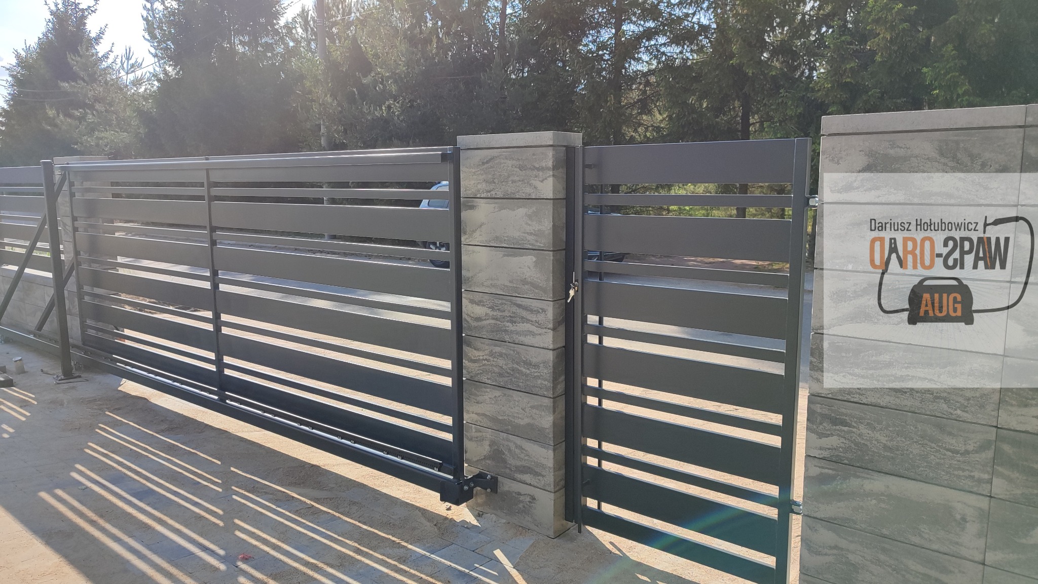Nowoczesna, przesuwna brama wjazdowa z poziomymi panelami w kolorze grafitowym, zamontowana na słupkach z szarego, warstwowego betonu, z widocznym logo firmy na jednym ze słupków.