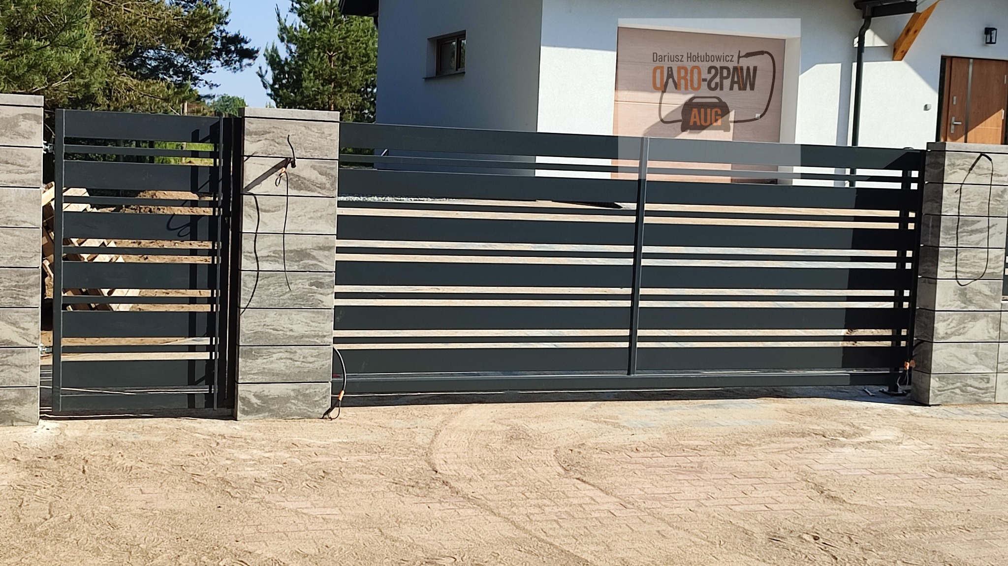 Nowoczesna, przesuwna brama wjazdowa z poziomymi panelami, osadzona na kamiennych słupkach, z widocznym logo firmy na garażu w tle.