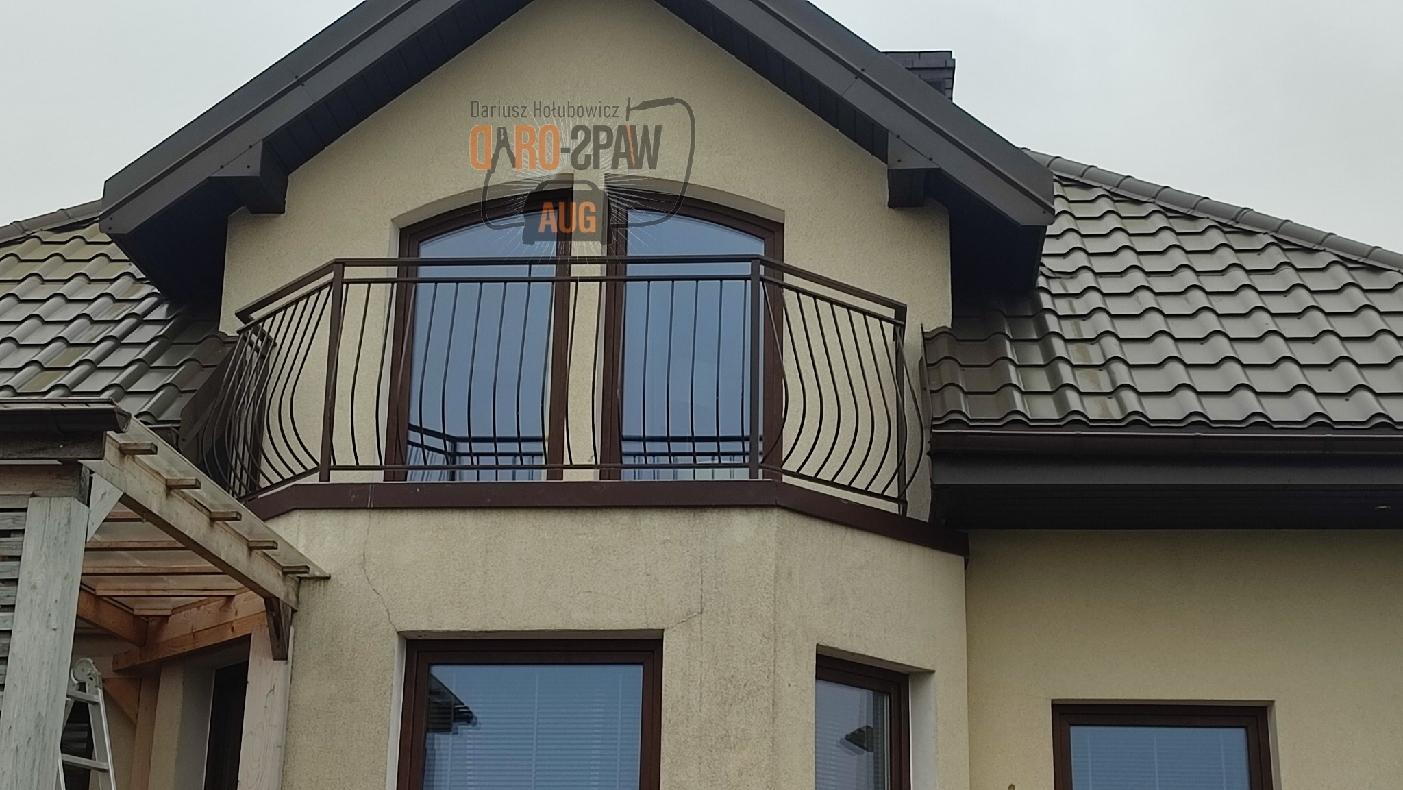 Elewacja domu z balkonem zabezpieczonym metalową balustradą o dekoracyjnym, falistym wzorze, widoczna z poziomu gruntu.