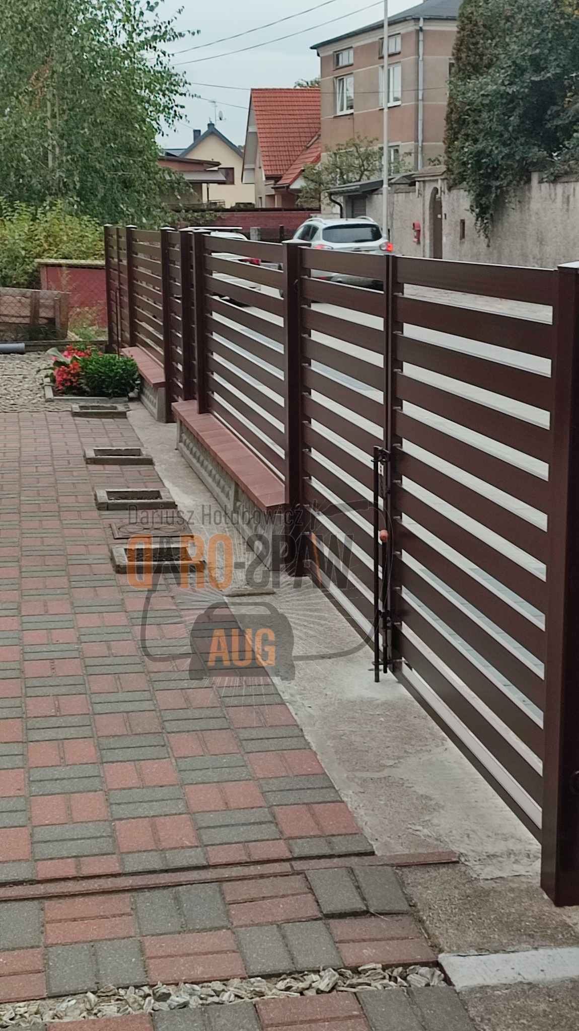 Nowoczesne, brązowe ogrodzenie posesji z poziomymi, białymi panelami, widok na furtkę i kostkę brukową na podjeździe.