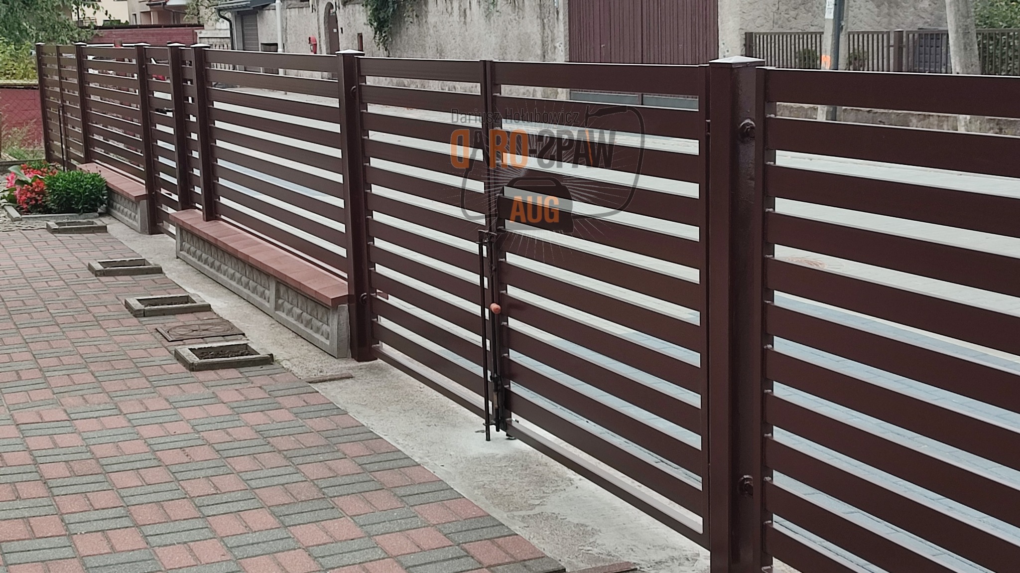 Brązowa brama wjazdowa z poziomymi panelami i prostym zamkiem, widok z zewnątrz na tle brukowanej ścieżki i zieleni.