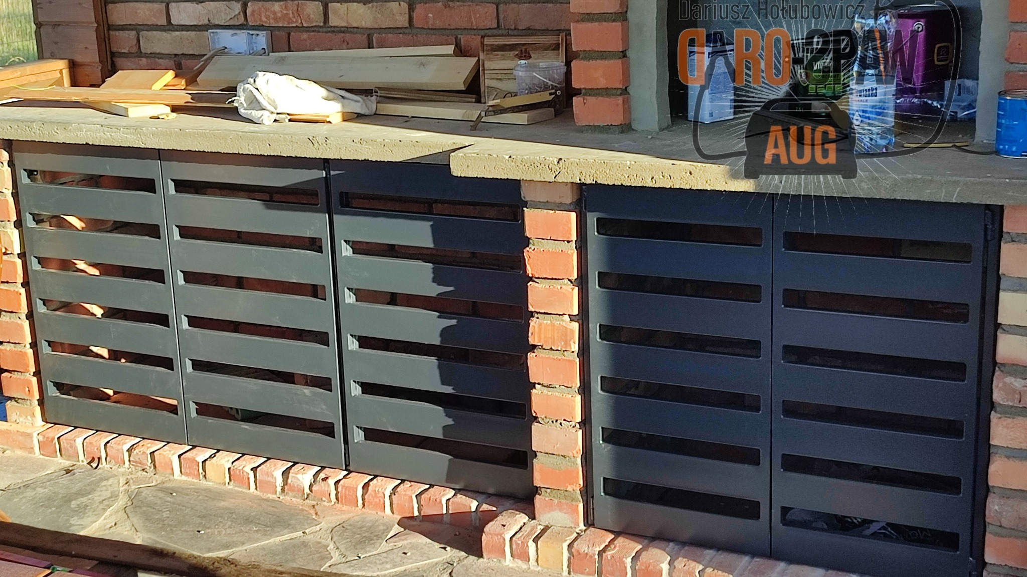 Zabudowa grilla z metalowymi, poziomymi lamelami w kolorze grafitowym, osadzona w ceglanej konstrukcji, widoczne narzędzia i materiały budowlane na blacie.
