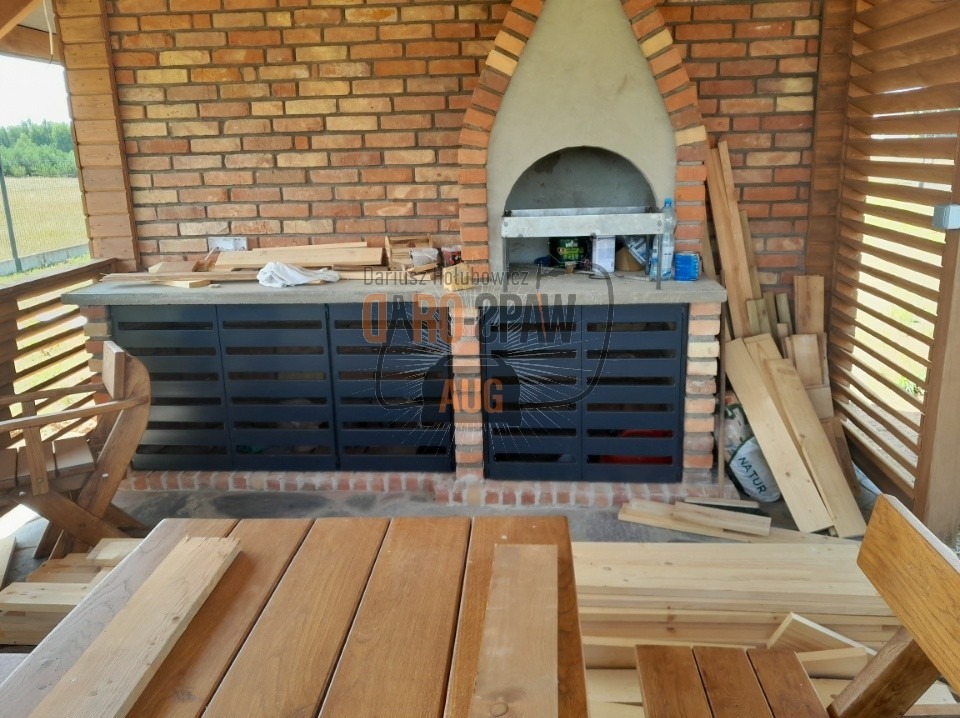 Zadaszona kuchnia letnia w trakcie budowy, z grillem murowanym z cegły i szafkami z metalowymi frontami z poziomymi szczelinami, widoczne deski i materiały budowlane.