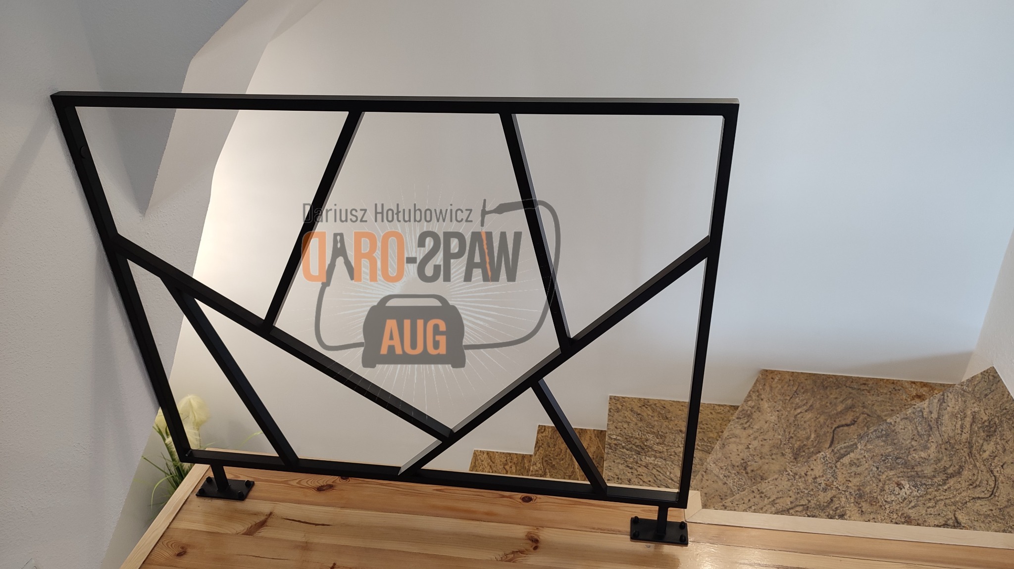 Czarna, metalowa balustrada schodowa o geometrycznym wzorze, zamontowana na drewnianym podeście, widok z góry na marmurowe schody, z delikatnym logotypem firmy w tle.