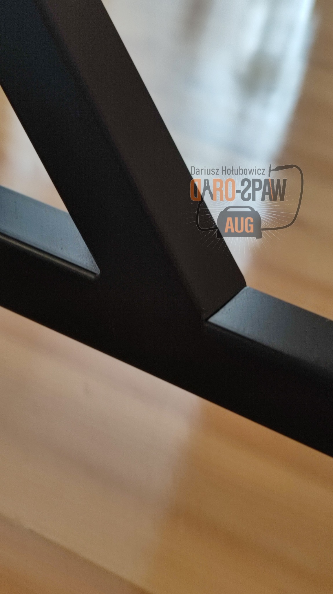 Szczegółowe ujęcie spawu na czarnej, metalowej konstrukcji, widoczny fragment logo firmy w tle, na drewnianej podłodze.