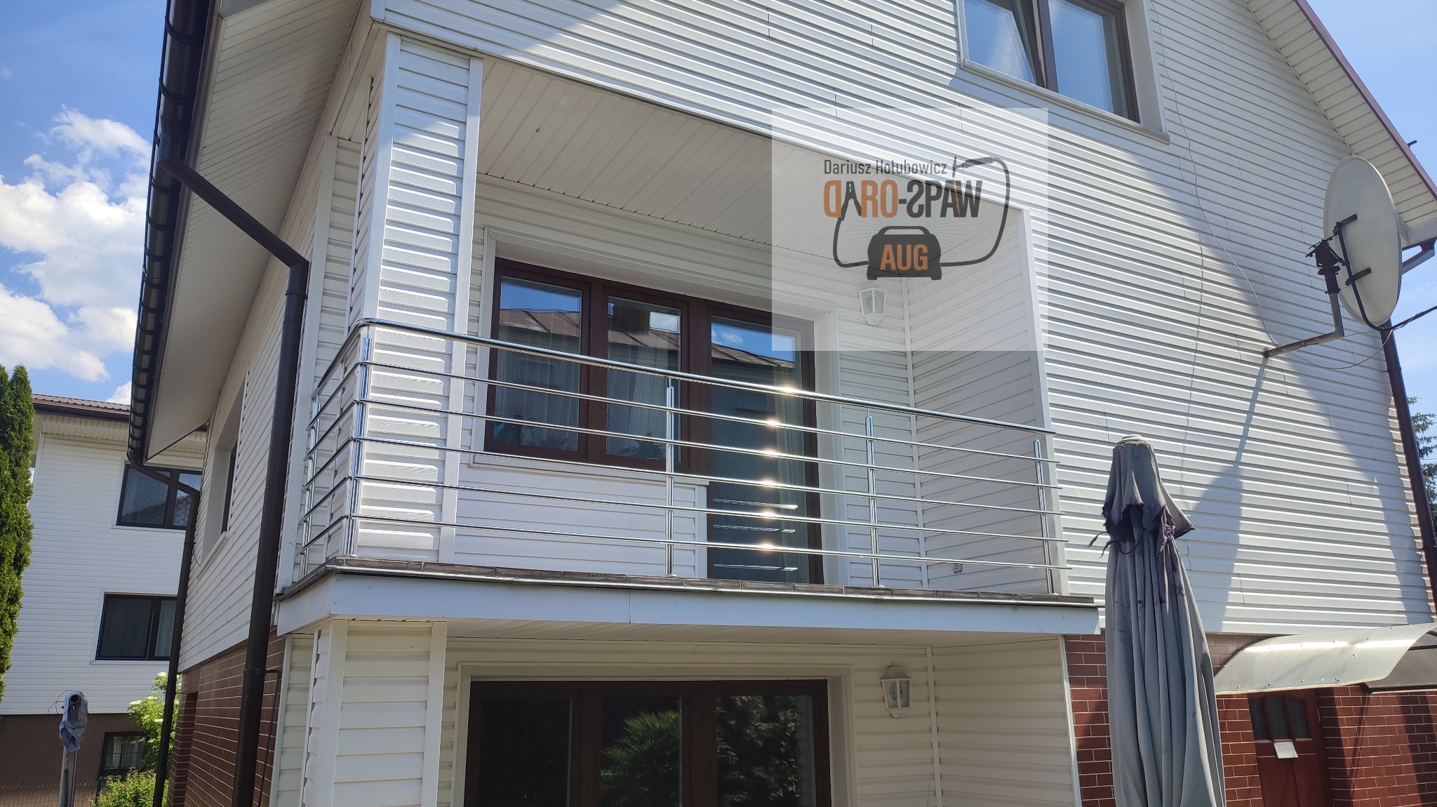 Biały dom z balkonem zabezpieczonym metalową balustradą z poziomych rurek, na elewacji logo firmy.