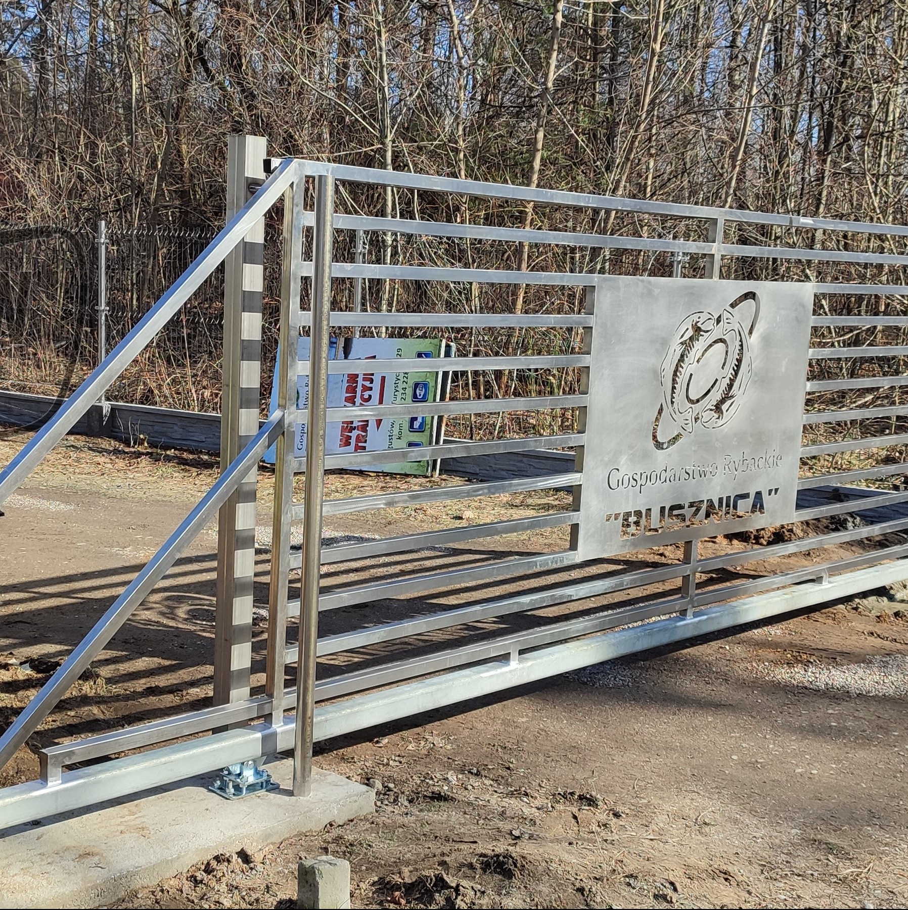Przesuwna brama wjazdowa z metalowymi poprzeczkami i ozdobnym panelem z wyciętym logo Gospodarstwa Rybackiego 'Bliznica', widoczny mechanizm jezdny i tło z drzewami.