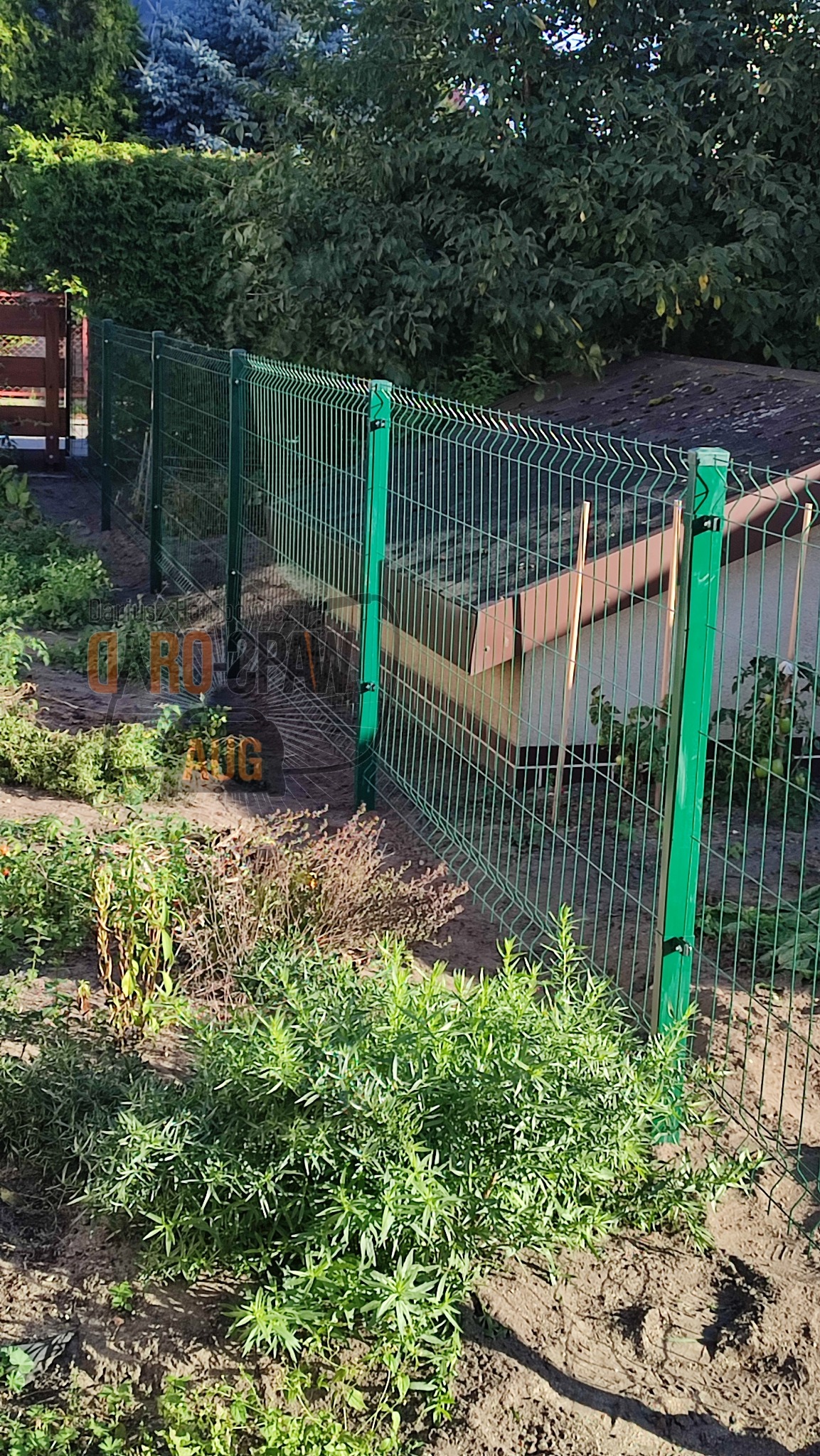 Zielone ogrodzenie panelowe z widoczną konstrukcją wsporczą, otaczające ogród z roślinnością i budynkiem w tle.