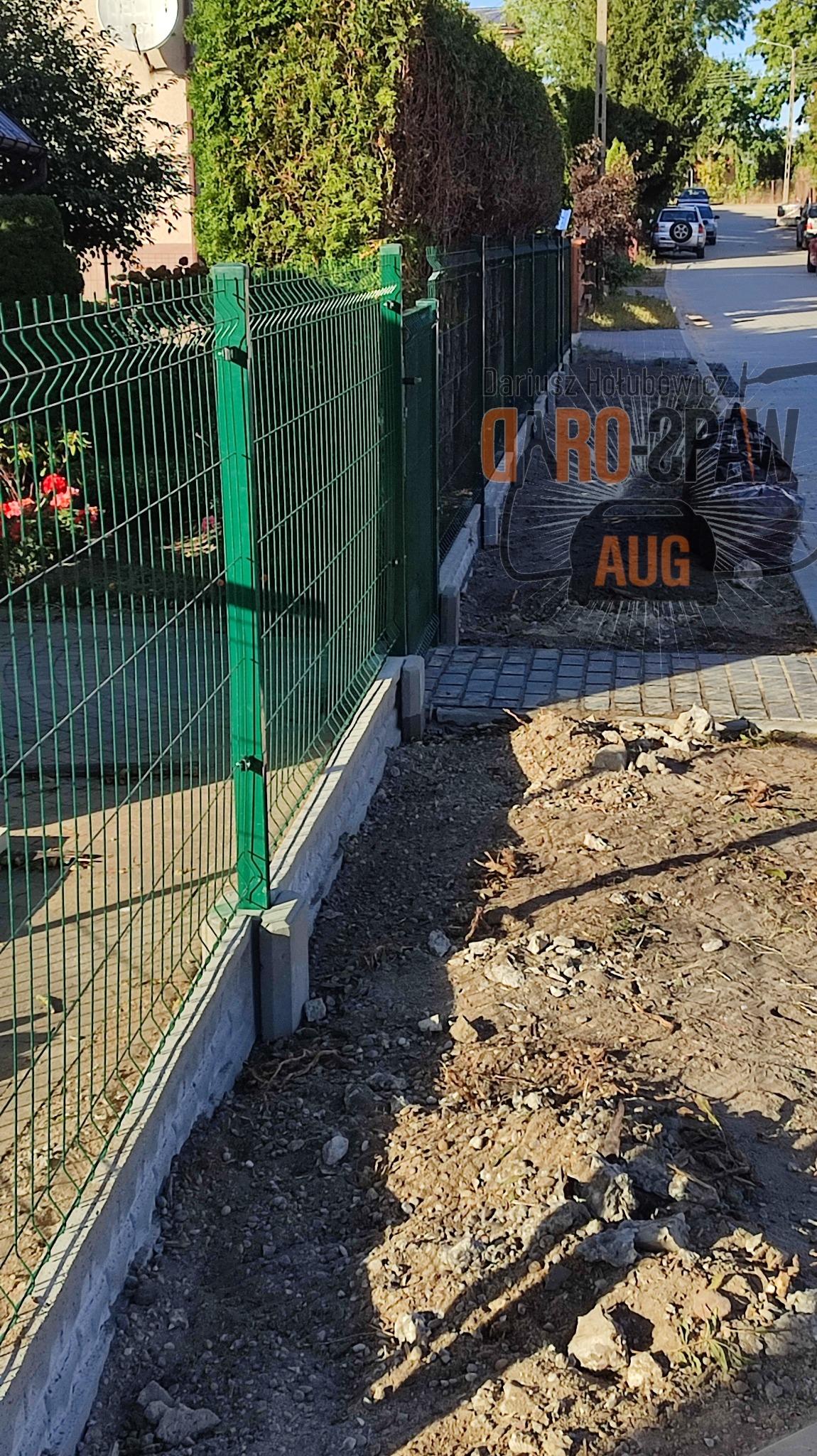 Fragment nowo zamontowanego, zielonego ogrodzenia panelowego z podmurówką betonową, widok z boku na nierówny teren przydomowy.