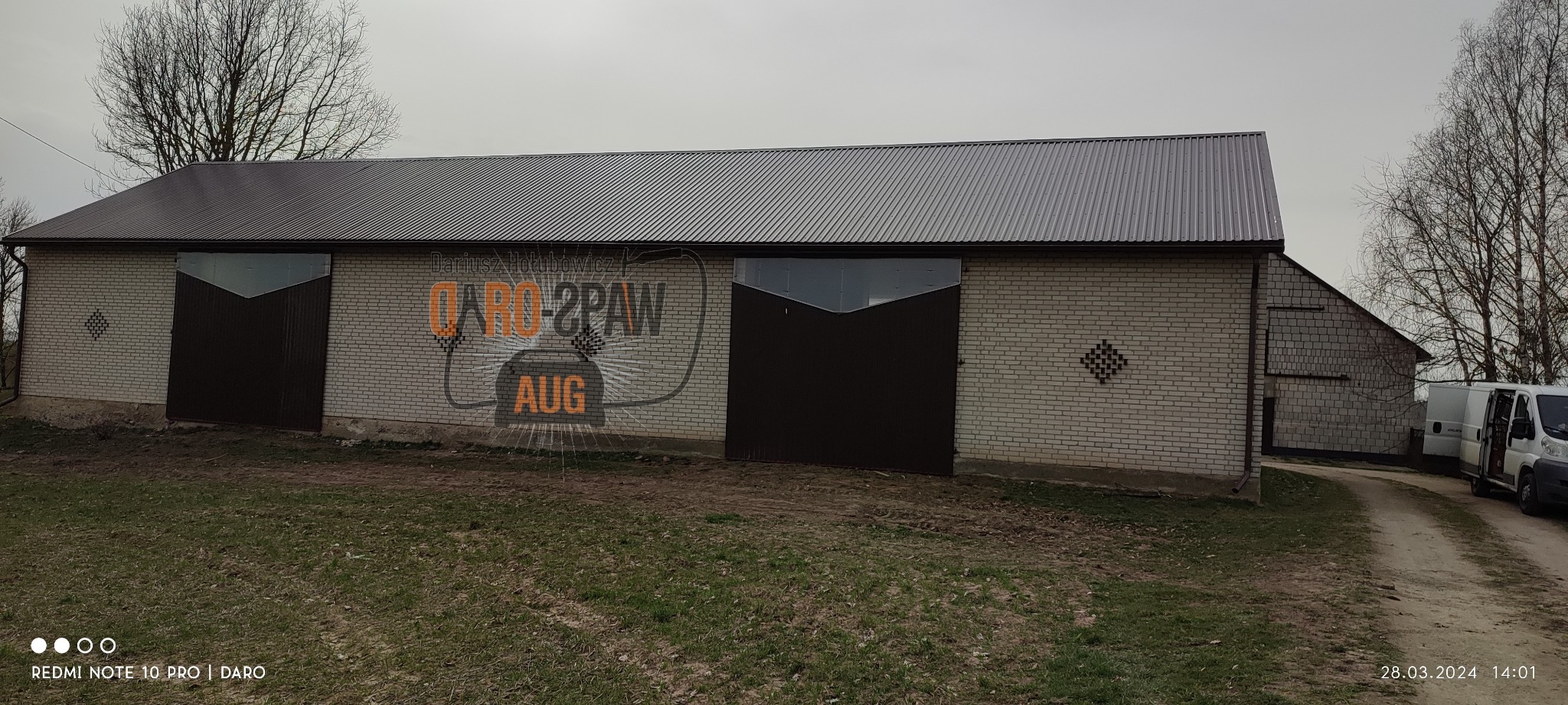 Duże, przesuwne, ciemne bramy wjazdowe do murowanego budynku gospodarczego z metalowym dachem. Na ścianie budynku widoczne logo firmy.