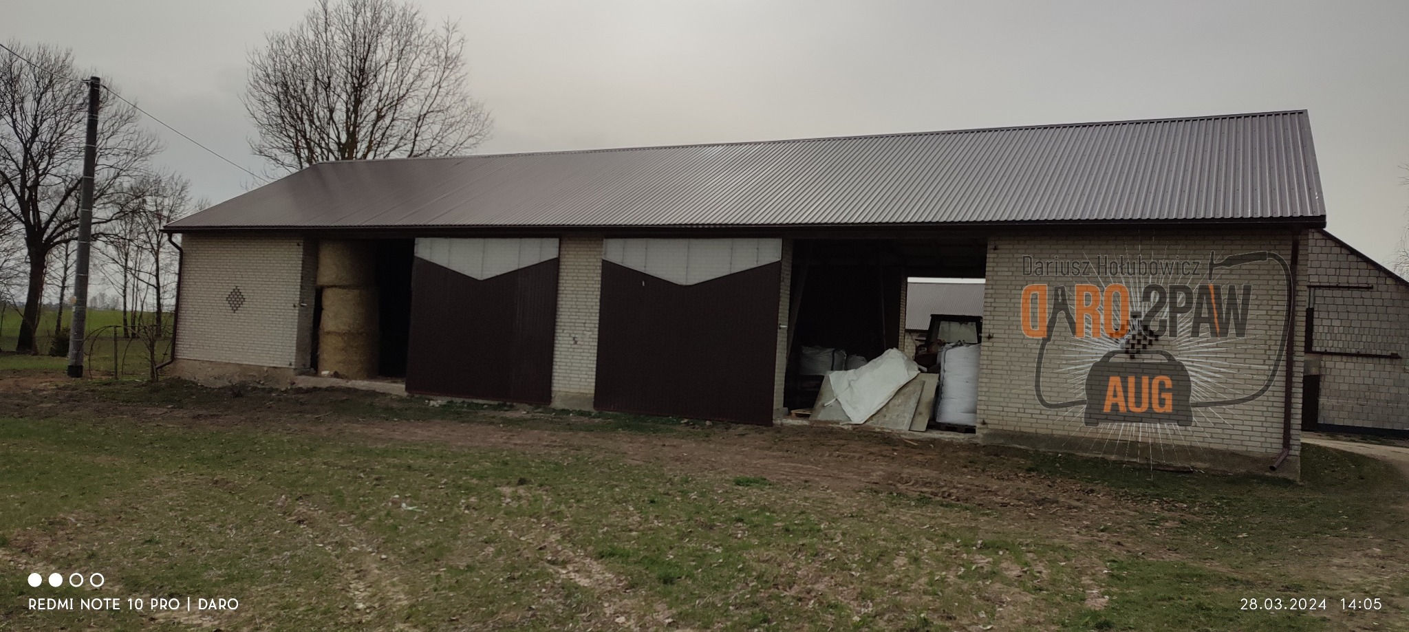 Brązowy blaszany dach i przesuwne dwuskrzydłowe wrota do murowanego budynku gospodarczego z widocznym logo firmy na ścianie.