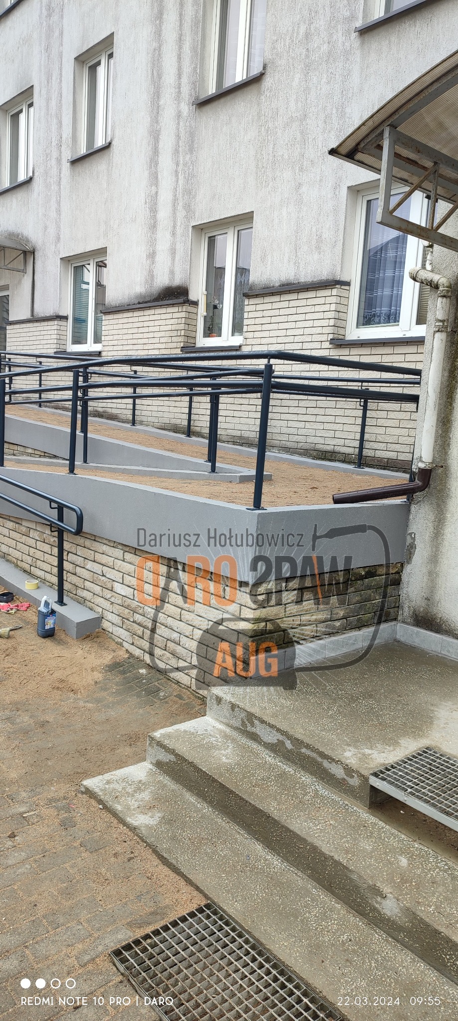 Zewnętrzna rampa dla niepełnosprawnych z czarną metalową balustradą, prowadząca do budynku z ceglaną elewacją, widok z dołu na schody wejściowe.