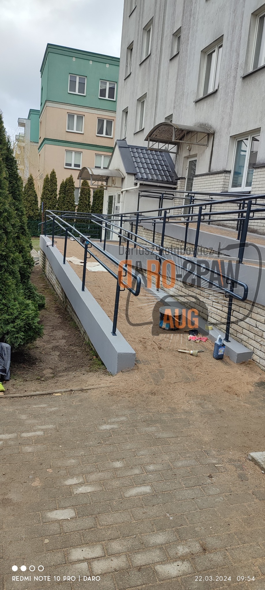 Rampa dla niepełnosprawnych z metalowymi poręczami przy budynku, widoczne narzędzia i puszka farby, teren po budowie.