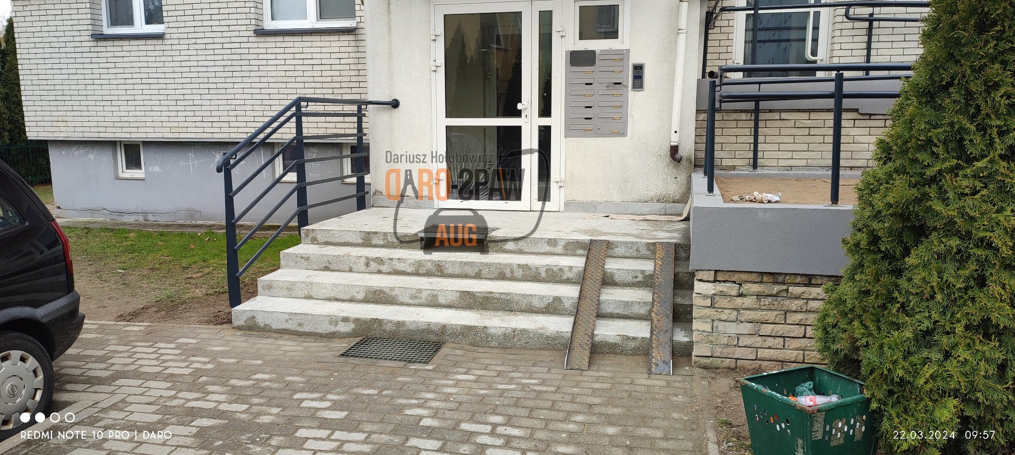 Metalowa balustrada schodowa przy wejściu do budynku, z rampą dla wózków inwalidzkich, widok z zewnątrz.