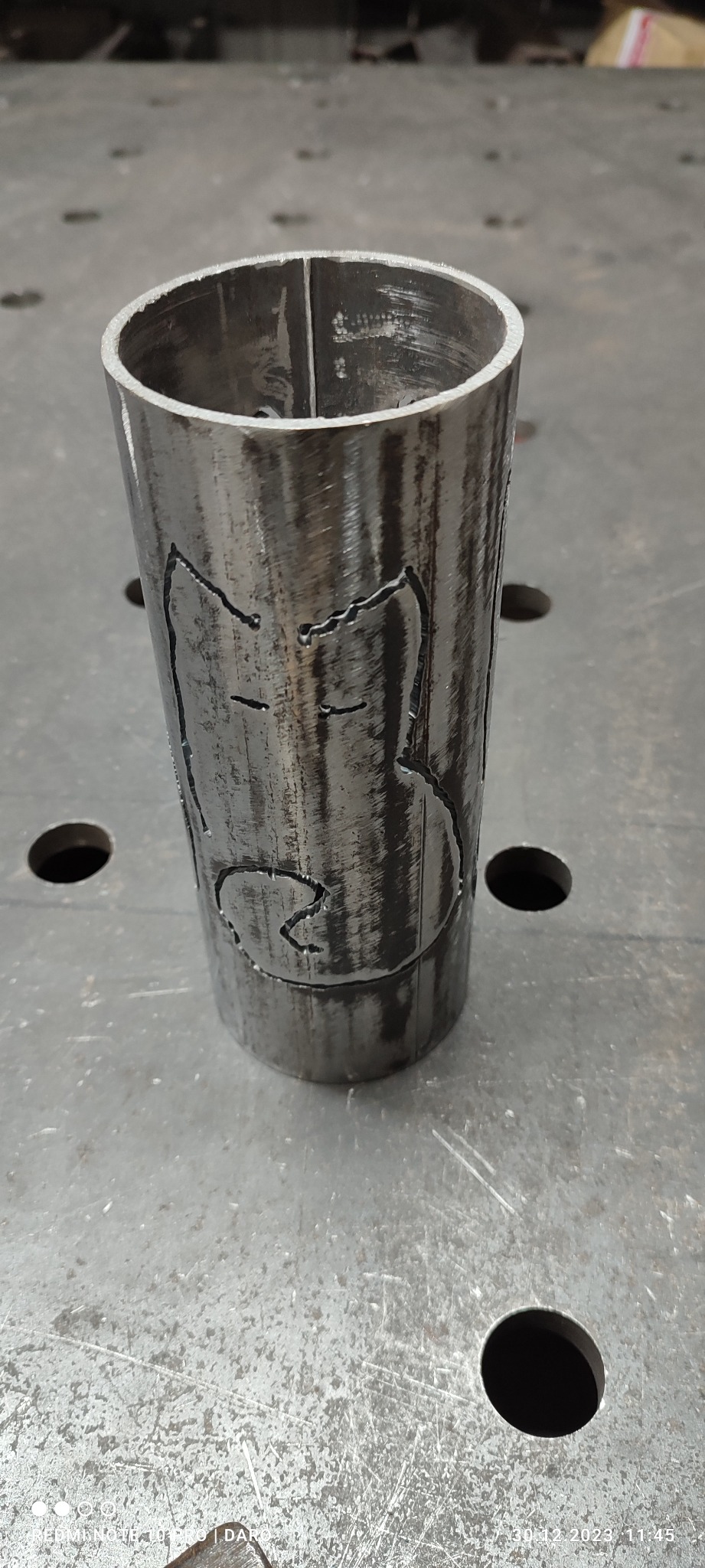 Metalowa rura z wyciętym laserowo wzorem przedstawiającym postać z kreskówki, leżąca na metalowym stole warsztatowym.