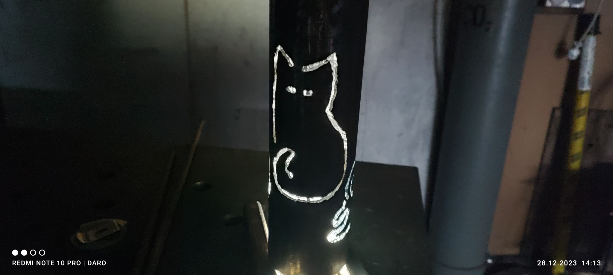 Metalowa rura z wyciętym wzorem kota, oświetlona od wewnątrz, widoczna butla z gazem w tle.