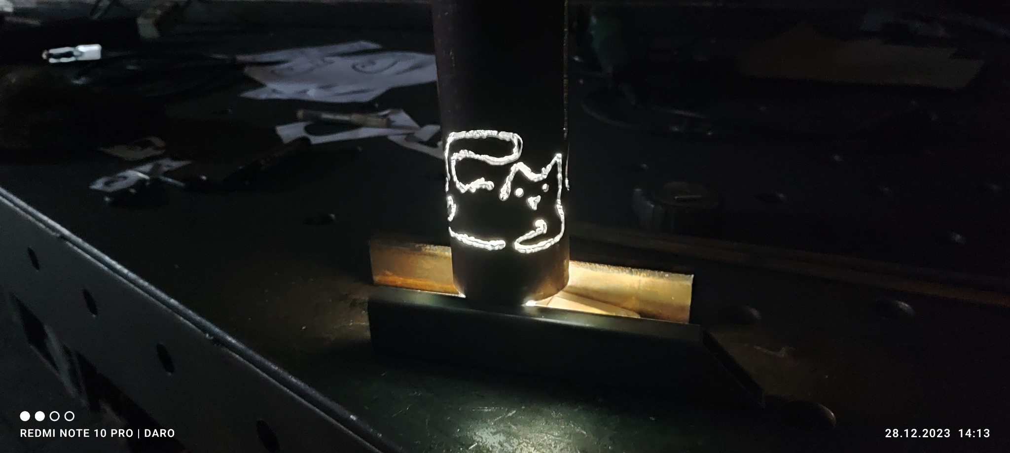 Metalowy cylinder z wyciętym laserowo wzorem kota, umieszczony na metalowej powierzchni warsztatowej.