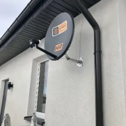 Szara antena satelitarna 'Cyfrowy Polsat HD' zamontowana na elewacji jasnego domu jednorodzinnego, widoczna drabina aluminiowa i rynna spustowa.