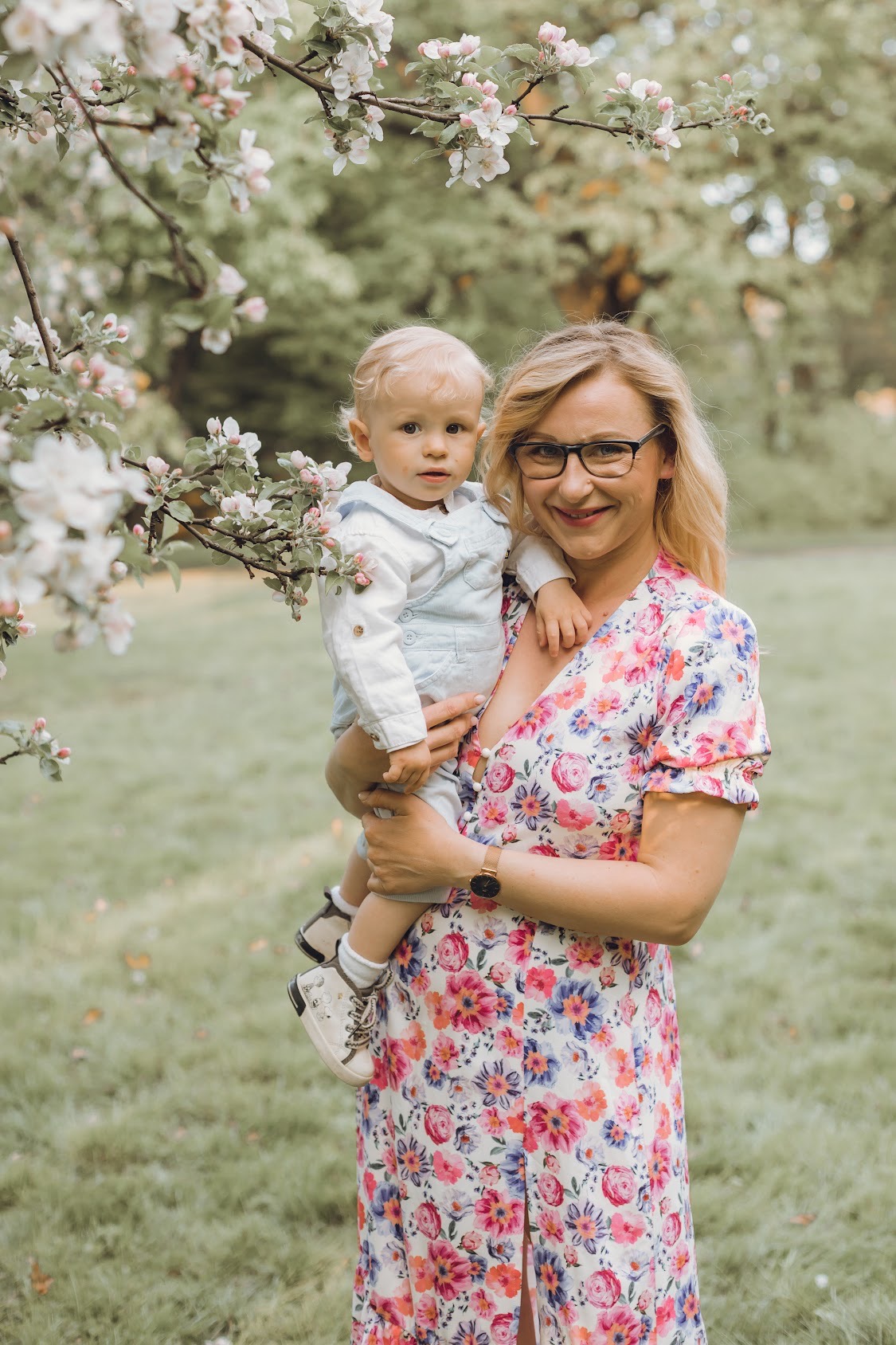 Uśmiechnięta blondynka w okularach trzyma na rękach małego chłopca na tle kwitnącego drzewa i zielonego trawnika.