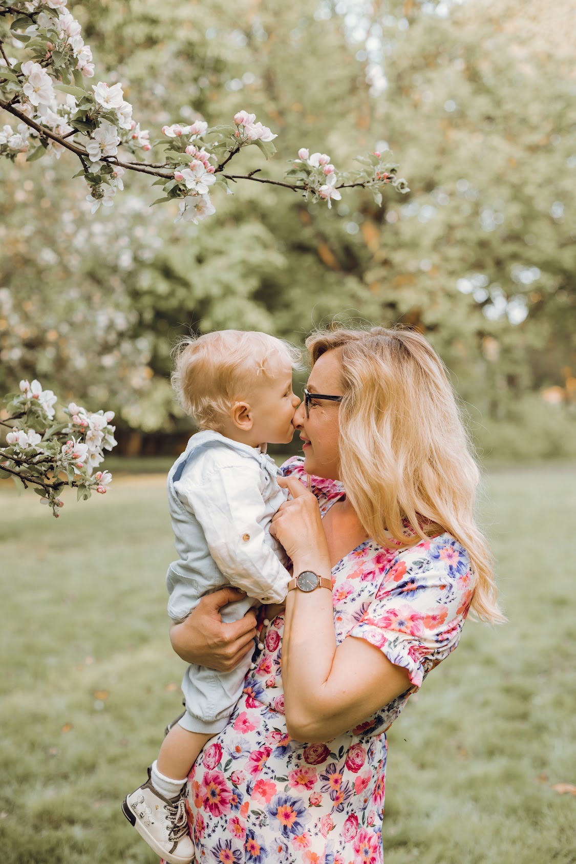 Matka w kwiecistej sukience trzyma na rękach blondwłosego synka, dotykając się nosami na tle kwitnących gałęzi drzewa.