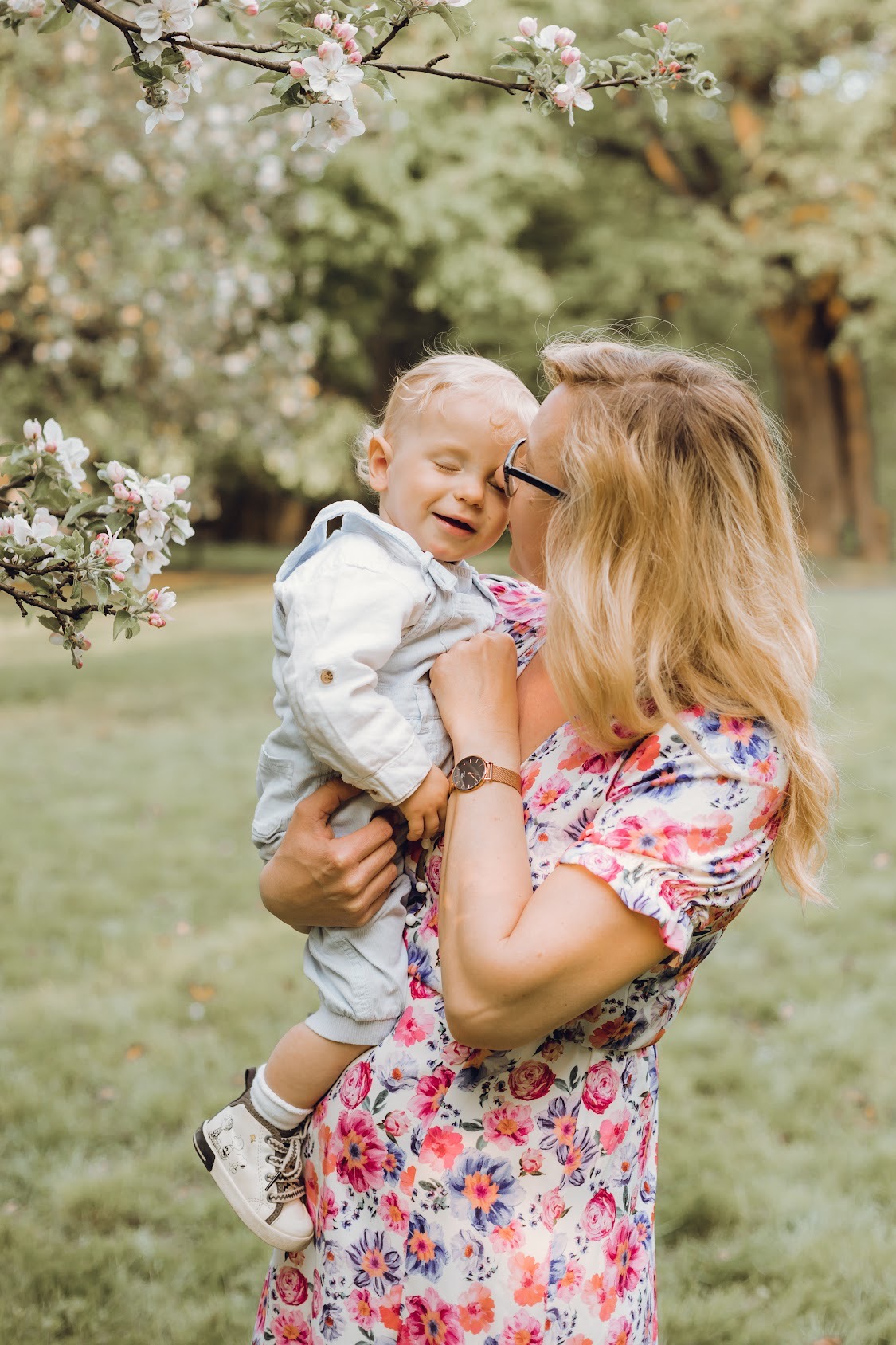 Matka w kwiecistej sukience trzyma uśmiechniętego blondwłosego synka na tle kwitnącego drzewa.