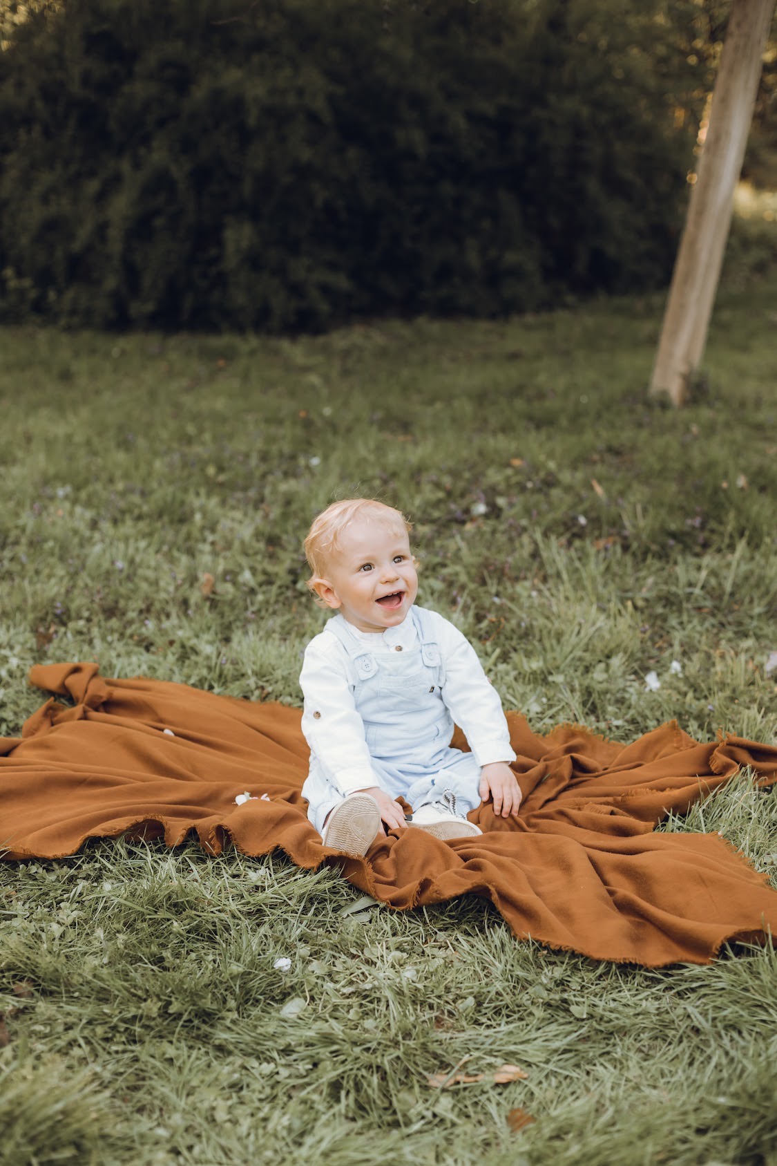 Radosny blondwłosy chłopiec w błękitnych ogrodniczkach siedzi na brązowym kocu na trawie, uśmiechając się szeroko w kierunku światła.