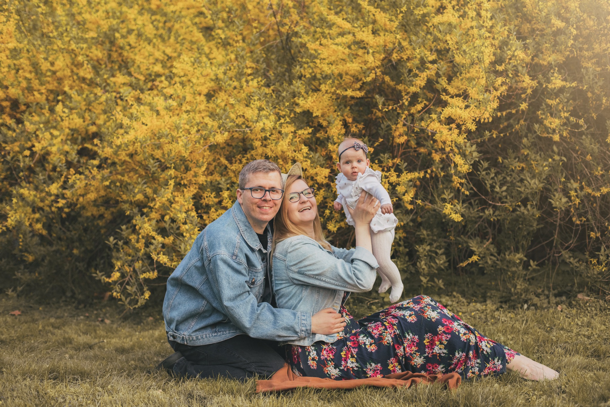 Szczęśliwa rodzina z małym dzieckiem na tle kwitnącego, żółtego krzewu podczas sesji zdjęciowej w Warszawie, mama trzyma córeczkę w górze.