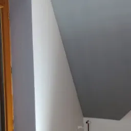 Pomalowane szarą farbą poddasze ze skośnym sufitem, widoczne rury i trzy białe haczyki na ścianie.