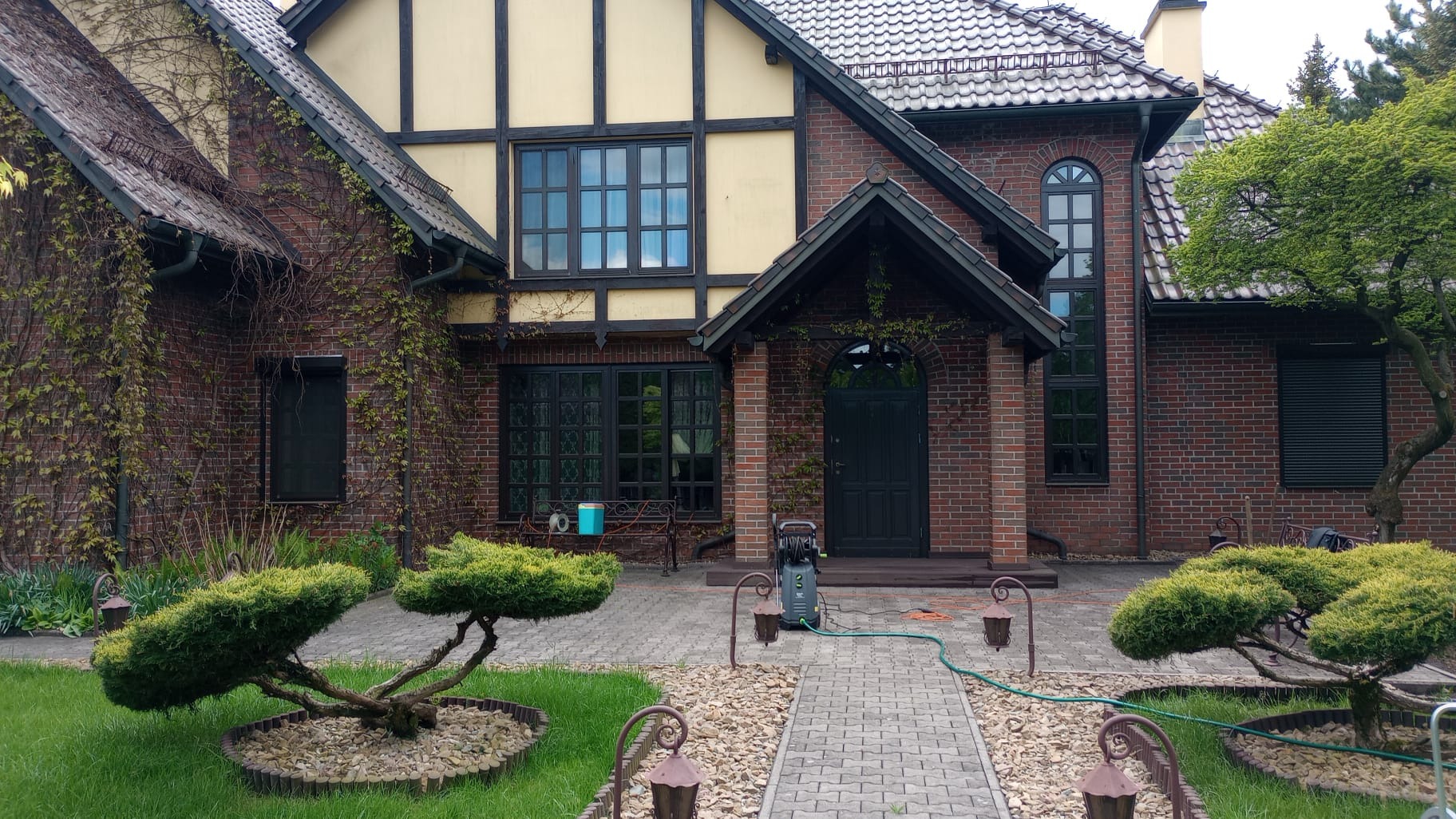 Czyszczenie elewacji domu z czerwonej cegły za pomocą myjki ciśnieniowej, widoczne zarośnięte pnącza na ścianach i zadbany ogród z ozdobnymi krzewami.