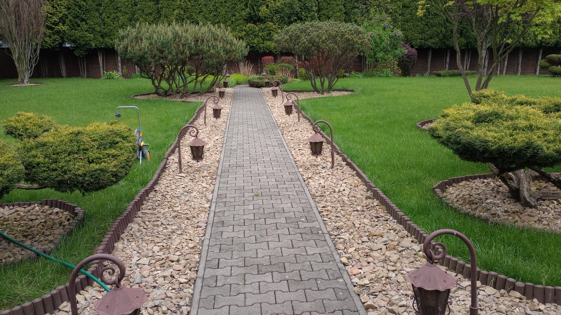 Uporządkowany ogród z trawnikiem, ścieżką z kostki brukowej i lampami ogrodowymi w stylu retro, otoczoną kamieniami i niskimi krzewami