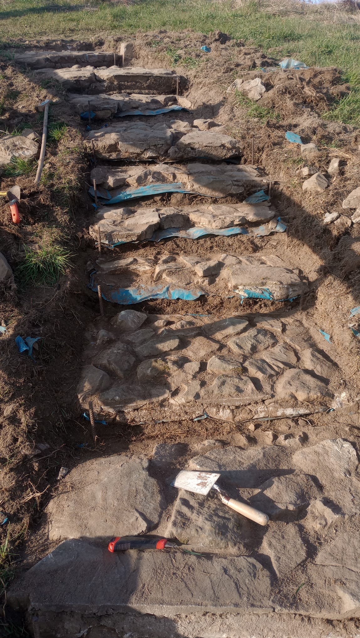 Budowa kamiennych schodów w ogrodzie, widoczne narzędzia: szpachelka i nóż monterski, niebieska folia pod kamieniami.
