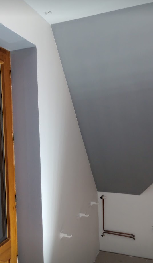 Pomalowane szarą farbą poddasze ze skośnym sufitem, widoczne rury i trzy białe haczyki na ścianie.