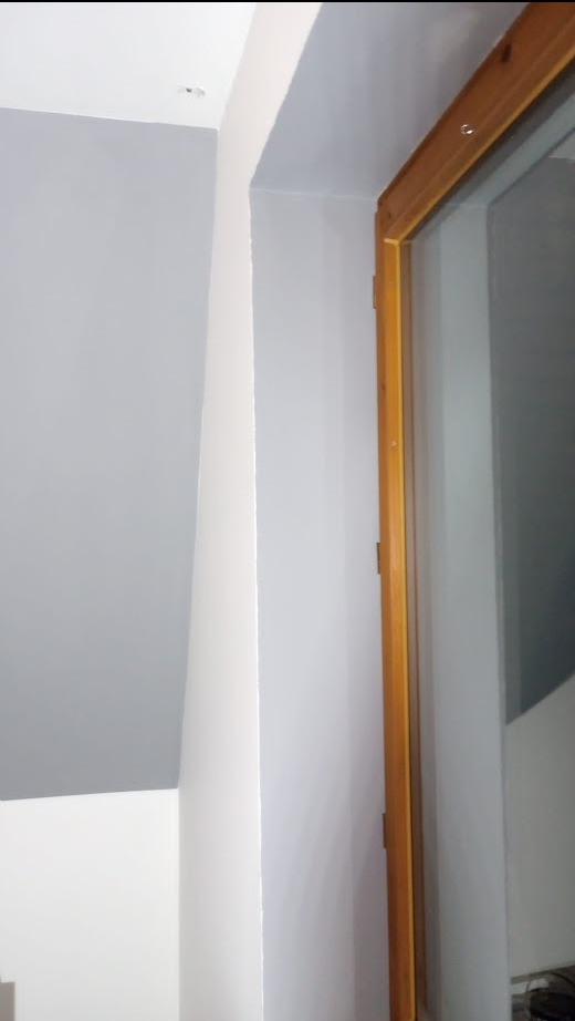 Świeżo pomalowane wnętrze z szarą ścianą i białym sufitem, widoczna drewniana rama okienna po prawej stronie.