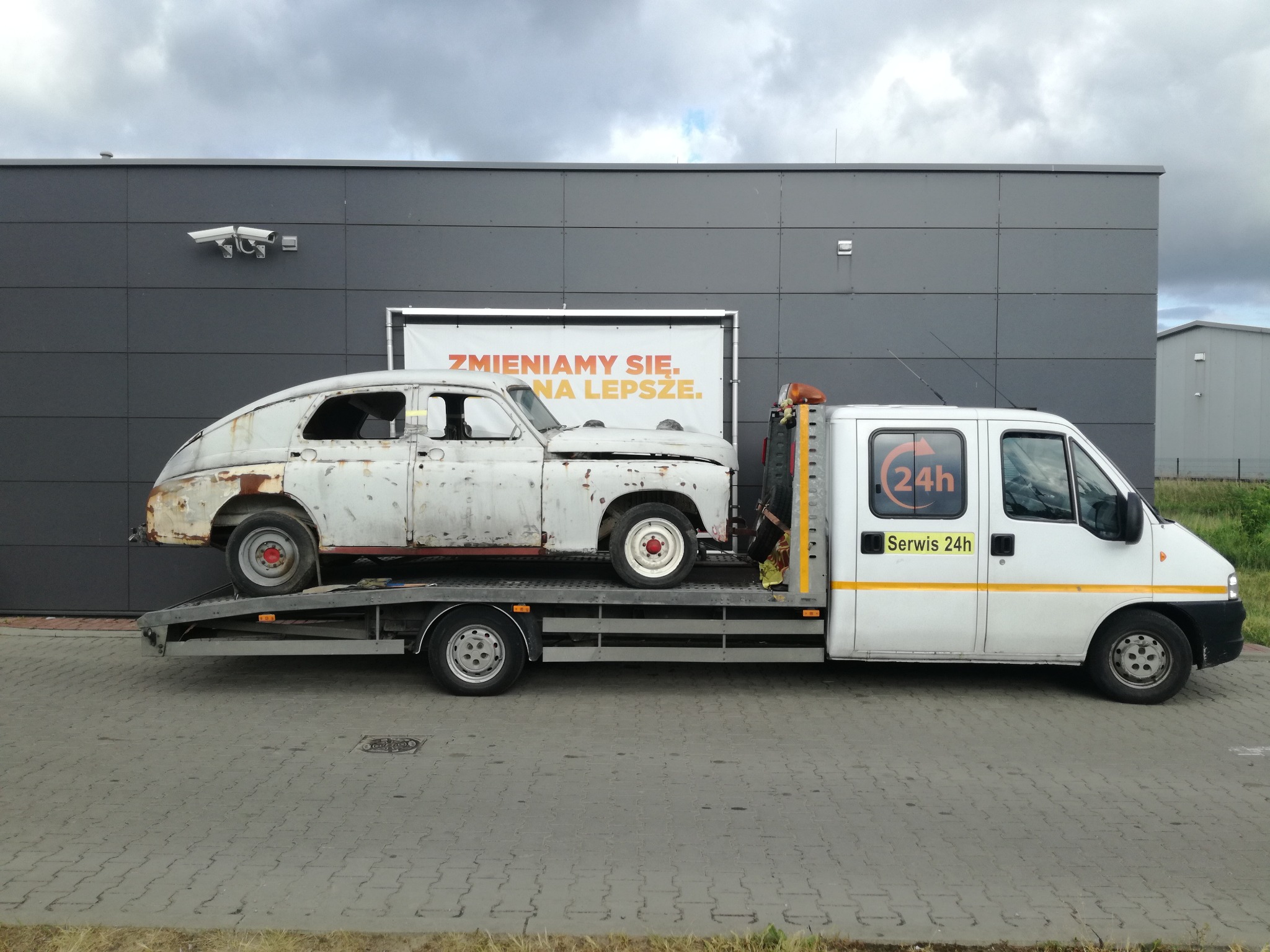 Laweta z białym busem przewozi skorodowany, zabytkowy samochód osobowy na tle szarej ściany budynku.