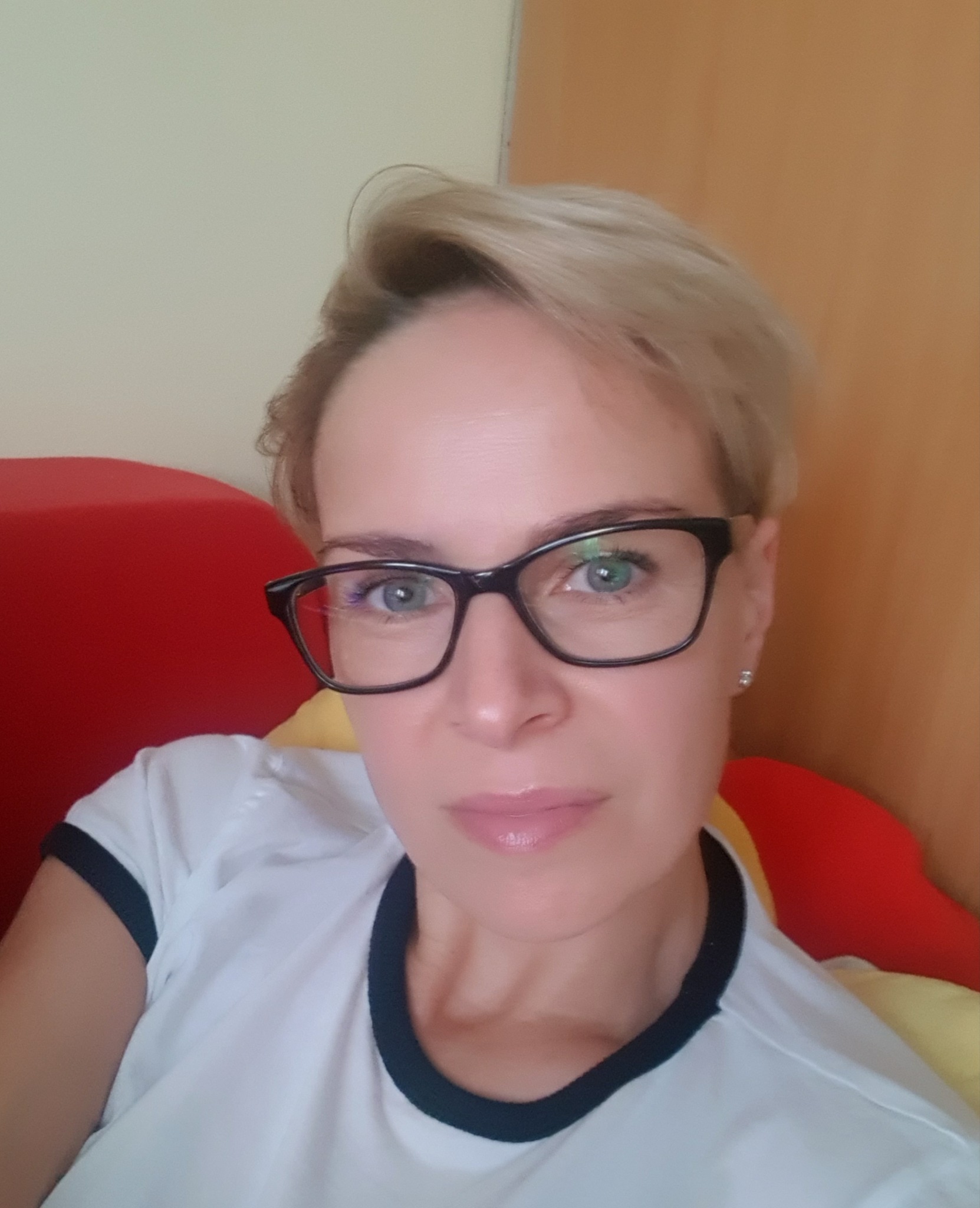 Portret kobiety z krótkimi blond włosami, w okularach z czarnymi oprawkami i białej koszulce z czarnym wykończeniem, na tle czerwonego i żółtego przedmiotu.