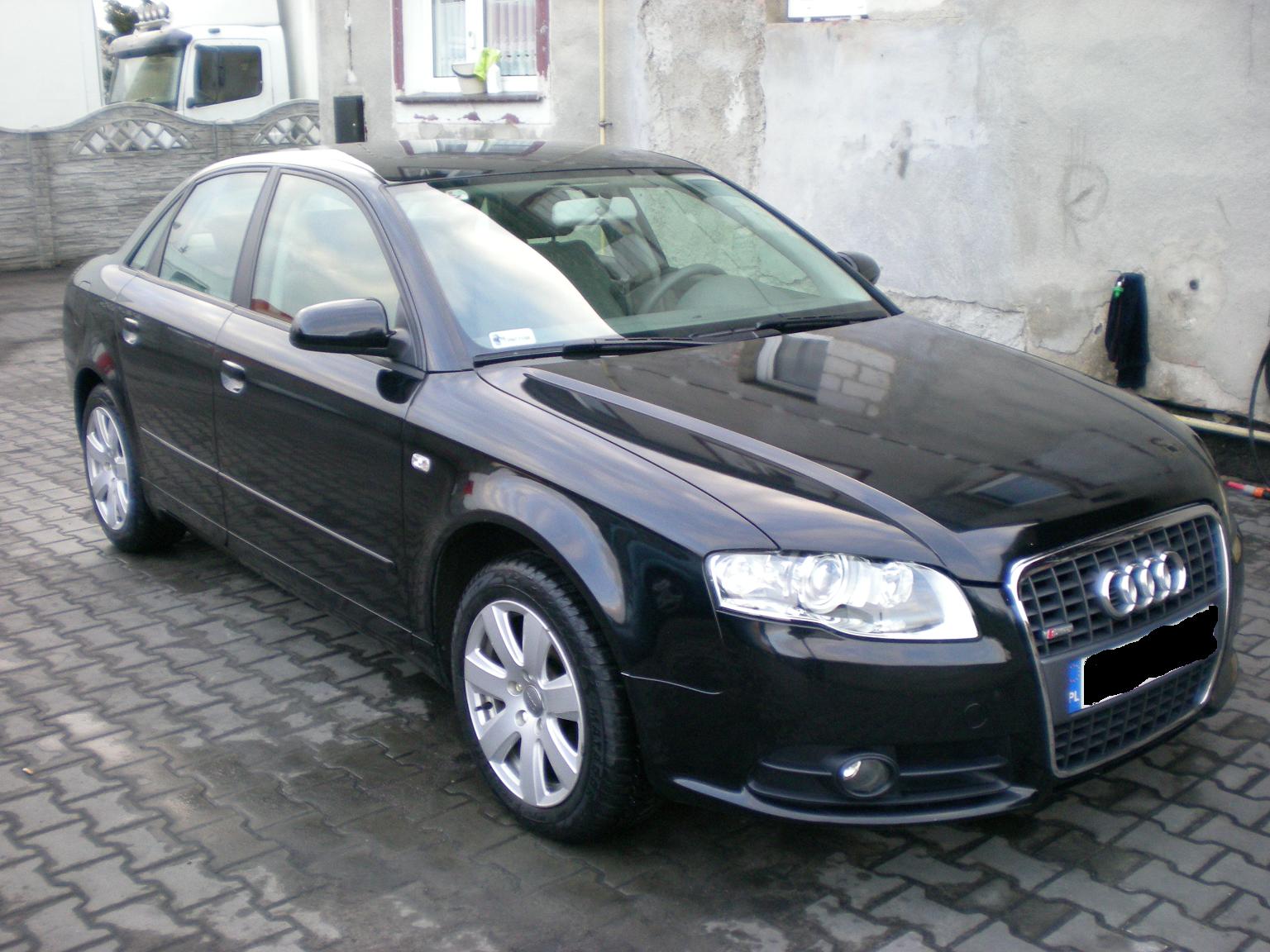 Czarny samochód Audi A4 1.9 TDI zaparkowany na mokrej kostce brukowej, z widocznym fragmentem białego busa w tle i szarą ścianą budynku.