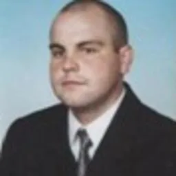 Portret mężczyzny w ciemnym garniturze i białej koszuli, na rozmytym, błękitnym tle.