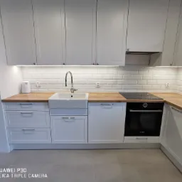 Jasna kuchnia z białymi szafkami i drewnianym blatem, widoczny zlew ceramiczny typu farmhouse, płyta indukcyjna i piekarnik Bosch.