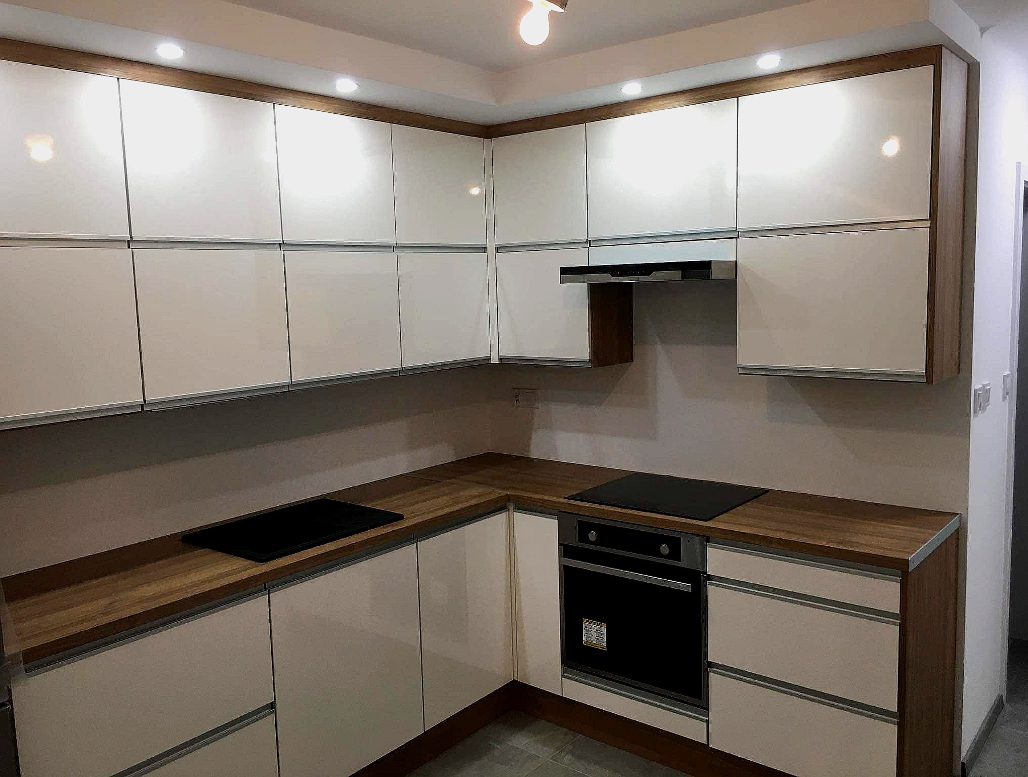 Nowoczesna kuchnia z białymi, lakierowanymi szafkami i blatem o ciepłym, drewnianym odcieniu, z wbudowaną płytą indukcyjną i piekarnikiem, oświetlona punktowymi lampami sufitowymi.