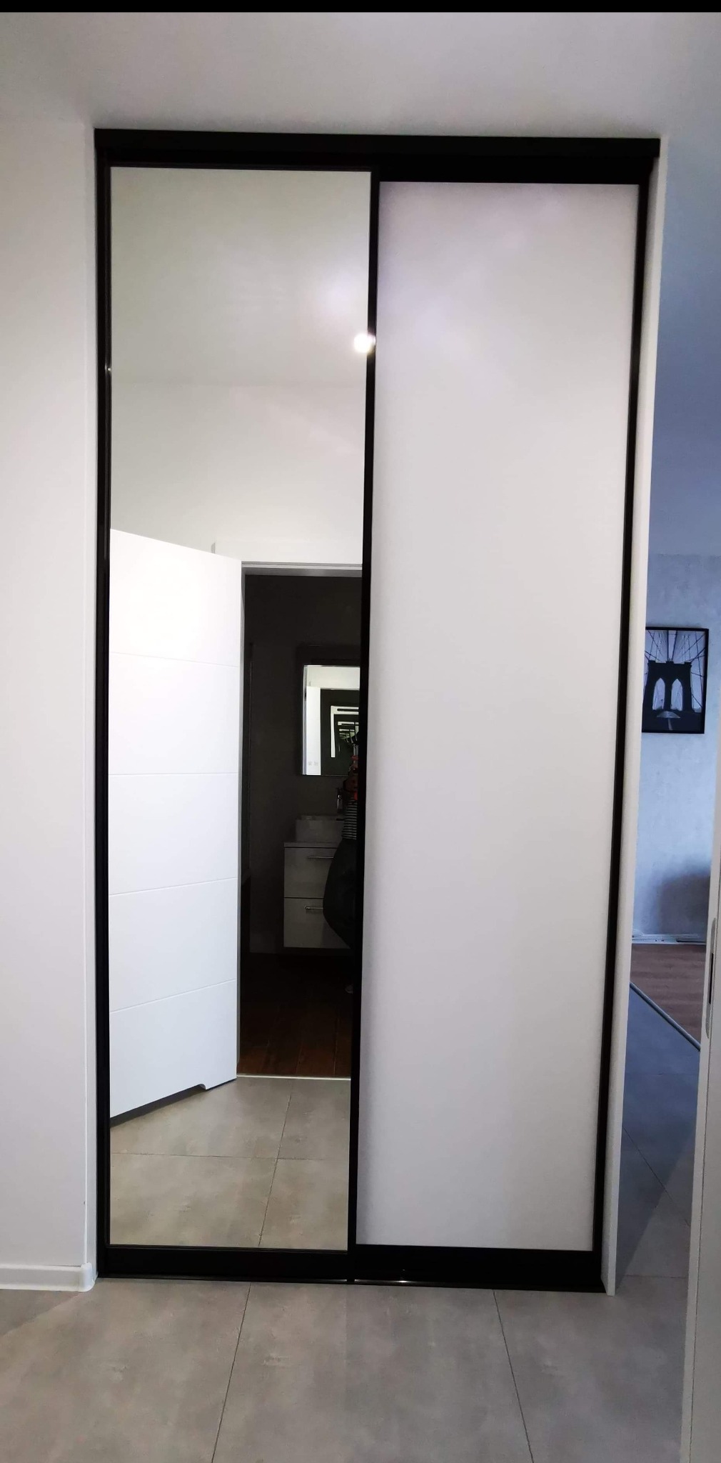 Nowoczesna szafa wnękowa z przesuwnymi drzwiami, czarne ramy, białe wypełnienie, widok na łazienkę z lustrem w tle.