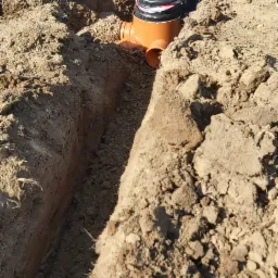 Pracownik w szarej kurtce pochyla się nad czarną studzienką kanalizacyjną z pomarańczowymi rurami, umieszczoną w wykopie. W tle widoczna łopata oparta o ziemię.