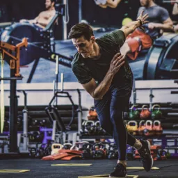 Mężczyzna w sportowym stroju wykonuje dynamiczne ćwiczenie koordynacyjne na tle wyposażonej siłowni we Wrocławiu, z widocznymi hantlami, kettlebells i plakatem z innymi sportowcami.