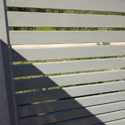 Fragment szarego ogrodzenia panelowego z poziomych profili, widok przez szczeliny na zieleń ogrodu, cień na murze w lewym górnym rogu.