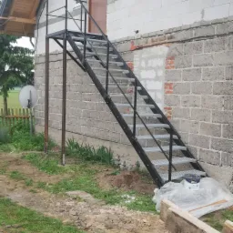 Zewnętrzne metalowe schody z platformą prowadzące do drzwi na piętrze budynku z szarego bloczka, widoczny fragment niezabezpieczonej ściany z cegły.