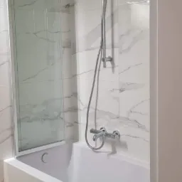 Nowoczesna łazienka z białą wanną, szklanym parawanem i marmurowymi płytkami na ścianach. Bateria wannowa chromowana z słuchawką prysznicową.